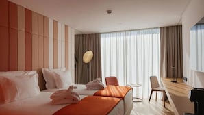 Habitación de hotel con dos camas, zona de estar con una silla, una mesita y un ventanal grande con cortinas.