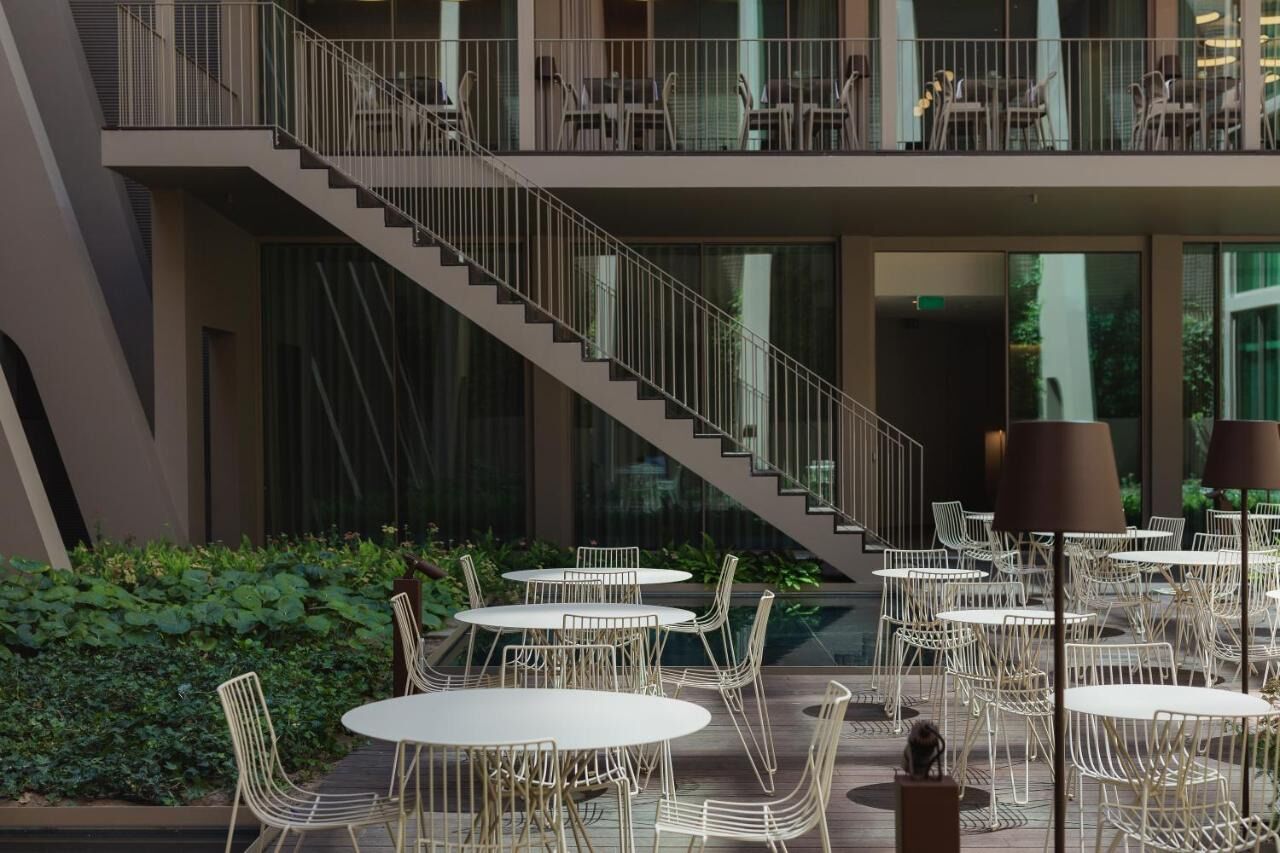 A modern outdoor dining area with round tables and chairs, a staircase, and a balcony with seating.