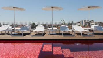 A poolside area with lounge chairs and umbrellas, a tiled pool, and a cityscape in the background.