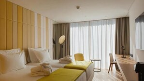 Hotellihuone, jossa on kaksi sänkyä, työpöytä, tuoli, pieni pöytä ja suuri ikkuna, jossa on verhot.