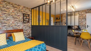 Camera da letto con parete in mattoni, un letto con biancheria a motivi decorativi, una scrivania con una sedia e una zona cucina visibile tramite un tramezzo.
