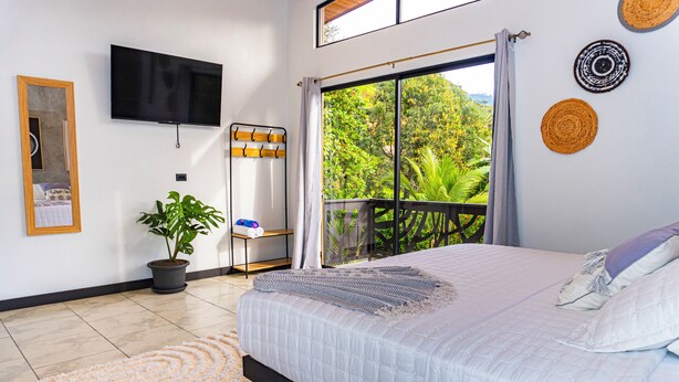 A bedroom with a large bed, a TV mounted on the wall, a potted plant, and a balcony with a view of greenery.