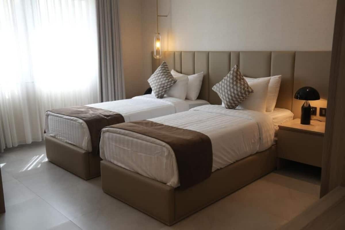 Habitación de hotel con dos camas, una mesita de noche con lámpara y una ventana con cortinas.