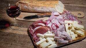 Eine Vorspeisenplatte mit verschiedenen Wurstwaren, Käsesorten und Essiggurken, dazu ein Brotlaib und ein Glas Rotwein auf einem Holztisch.