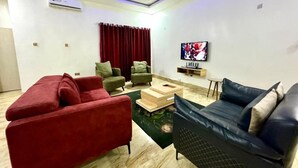 A modern living room with a red sofa, a blue sofa, and green armchairs. There is a flat-screen TV mounted on the wall and a wooden coffee table in the center. - Marcopolo Hotel and Suites  (Lekki)