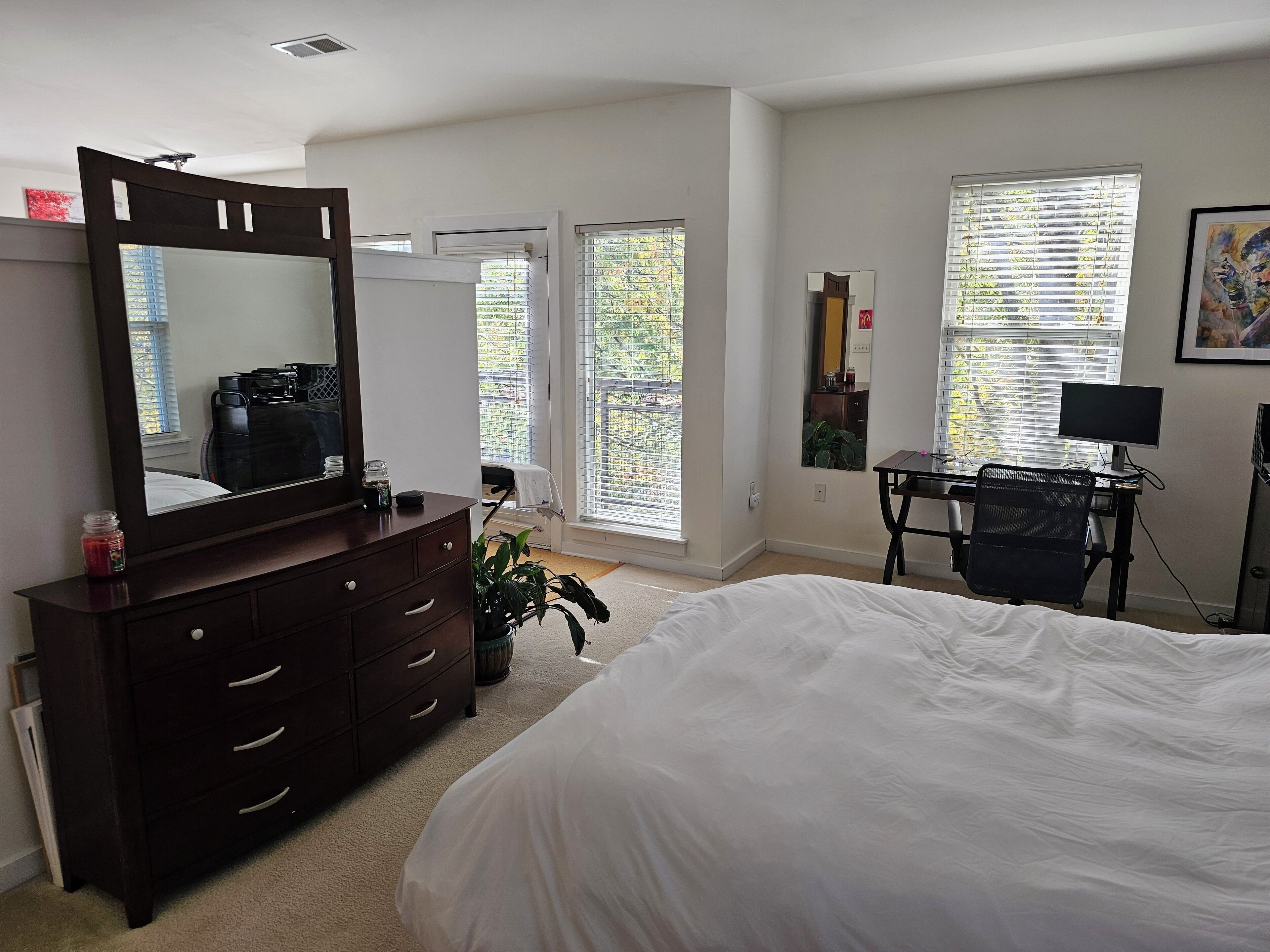 Une chambre à coucher avec un grand lit, une commode avec des tiroirs, un miroir, un bureau avec un ordinateur et une fenêtre avec des stores.