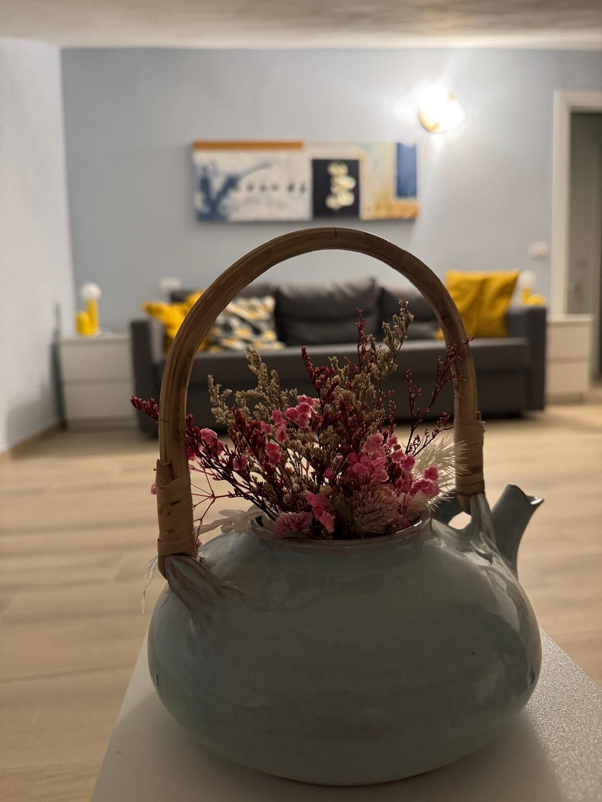 Une théière en céramique grise, dotée d’une anse en bambou et contenant un arrangement floral, est posée sur une surface blanche dans une pièce où se trouvent un canapé gris et un coussin jaune en arrière-plan.