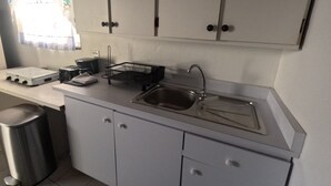 Ett kompakt kök med vita skåp, en diskho i rostfritt stål och ett fönster med blå gardiner.