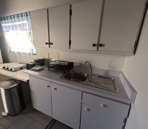 A compact kitchen with white cabinets, a stainless steel sink, and a window with blue curtains. - Cozy 1-bedroom apartment in beautiful Noord with AC (Noord)