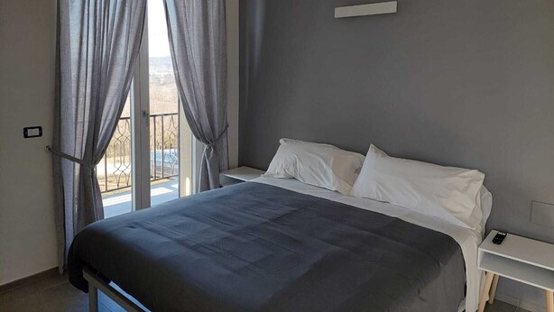Ein Schlafzimmer mit einem Bett, weißen Kissen, einem Nachttisch mit Fernbedienung und Blick ins Freie durch ein Fenster mit Vorhängen.