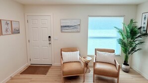 A room with a wooden floor, two leather chairs with white cushions, a small round table, a potted plant, a door with a keypad lock, and a window with blinds. - Shar’s Cottage - Close to Beach - Bradenton (Bradenton)