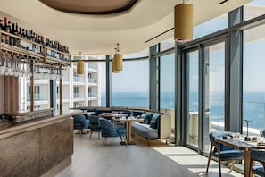 Ein moderner Barbereich mit Weinflaschen und Gläsern, eine Sitzecke mit Sofas und Tischen sowie große Fenster mit Blick auf das Meer.