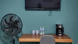 Una habitación con pared verde azulado, una mesa de madera, un ventilador negro, una silla gris, un televisor sobre la mesa y una cafetera en la mesa.