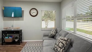 Un salon avec un canapé gris, un tapis à motifs, un téléviseur à écran plat, une horloge et une fenêtre avec des stores.