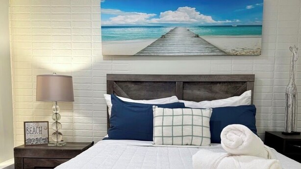 Un lit bien fait, recouvert d’une couette blanche, avec deux oreillers bleus et une serviette blanche enroulée. Un tableau représentant une plage avec une jetée est accroché au mur au-dessus du lit.