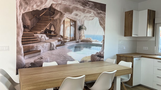 A modern kitchen with a dining area featuring a wooden table and white chairs, a stone wall mural depicting a mountainous landscape with a pool, and wooden cabinets. - Casa las Cordobesas is a charming rural lodging in the middle of nature. (La Guijarrosa (Córdoba))