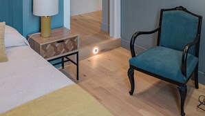 Ein Schlafzimmer mit einem Bett, einem Nachttisch mit Lampe, einem Sessel, einem runden Spiegel und einer Tür zu einem anderen Raum.