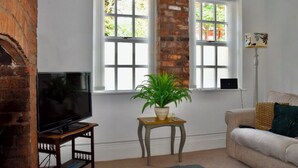 A living room with a TV on a wooden stand, a potted plant on a small table, a sofa with cushions, and a brick wall. - Coach house cottage by sefton park, built in 1850.  Entire house ! (Liverpool)