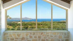 A room with a large window offering a view of a coastal landscape, stone walls, and a wooden ceiling. - Villa Athena (Apokoronas)