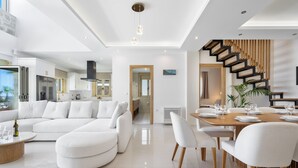 A modern living room with a sectional sofa, a dining area with a wooden table and chairs, and a staircase with a unique design. - Villa Athena (Apokoronas)