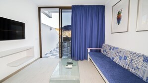 A modern living room with a glass coffee table, a sofa with patterned cushions, a flat-screen TV mounted on the wall, and a view of a cityscape through a sliding glass door. - Apartamentos Monte Verde (Mogán)