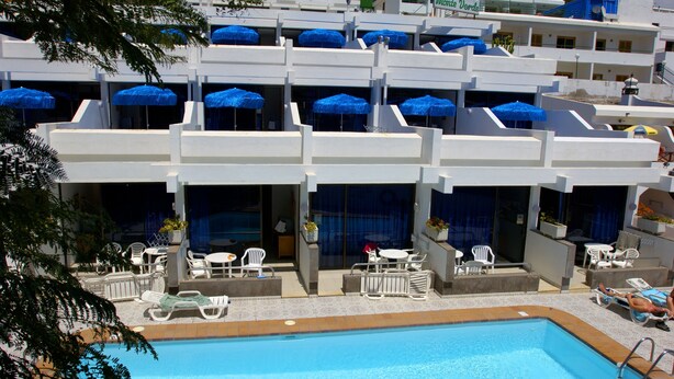A hotel with a swimming pool, outdoor seating, and blue umbrellas. - Apartamentos Monte Verde (Mogán)