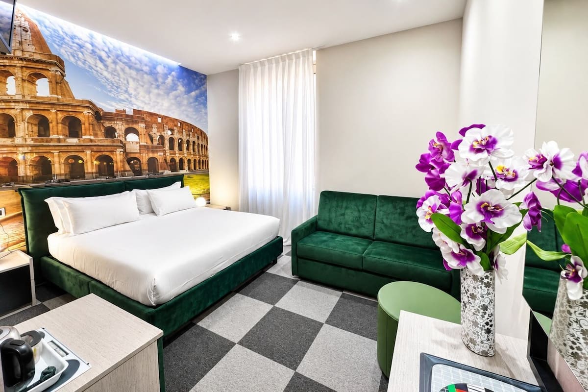 Quarto de hotel com cama, sofá verde, uma pequena mesa, um vaso com flores e um mural do Coliseu.