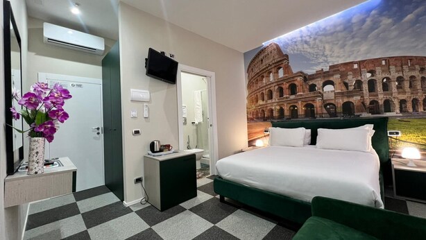 Una habitación de hotel con cama, mesita de noche, espejo, televisor montado en la pared, puerta de baño y un mural del Coliseo.