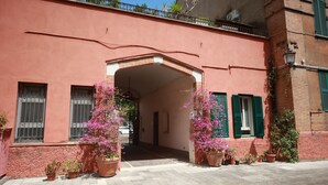 Un edificio con fachada rosa, contraventanas verdes y un arco central. Hay plantas en macetas y una entrada empaveda.