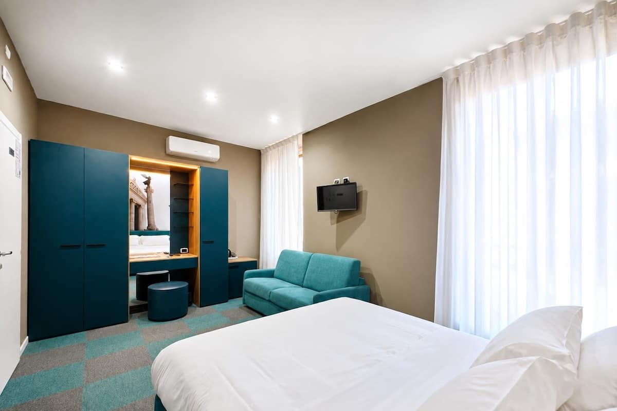 Quarto de hotel com cama, escrivaninha, cadeira, guarda-roupa e televisão.