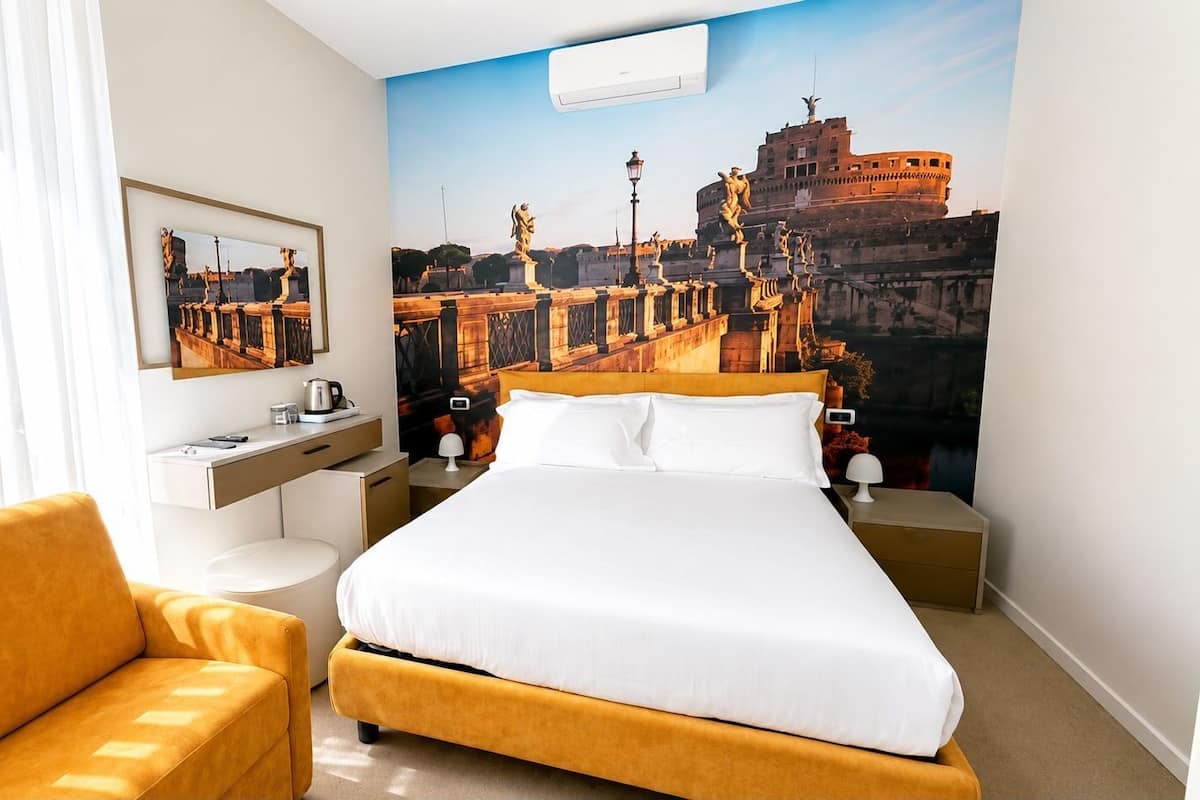 Quarto de hotel com cama, escrivaninha, cadeira e um grande mural de uma paisagem urbana histórica.