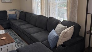 Een woonkamer met een donkergrijze hoekbank, een klein houten bijzettafeltje, een kamerkleed met dessin en een raam met dunne gordijnen.