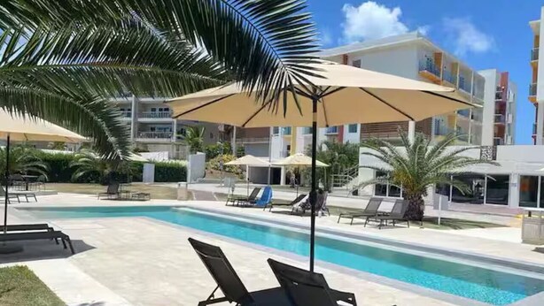 En swimmingpool med solstolar och parasoller, omgiven av palmer och moderna byggnader.