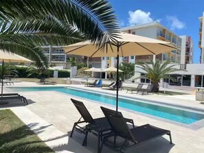 A swimming pool with lounge chairs and umbrellas, surrounded by palm trees and modern buildings. - Luxury Maho Entire Apt 1BR Ocean Runway View (Maho)