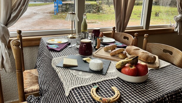 Ontbijttafel gedekt met eieren, brood, boter en fruit, met uitzicht op een landelijk landschap door grote ramen.