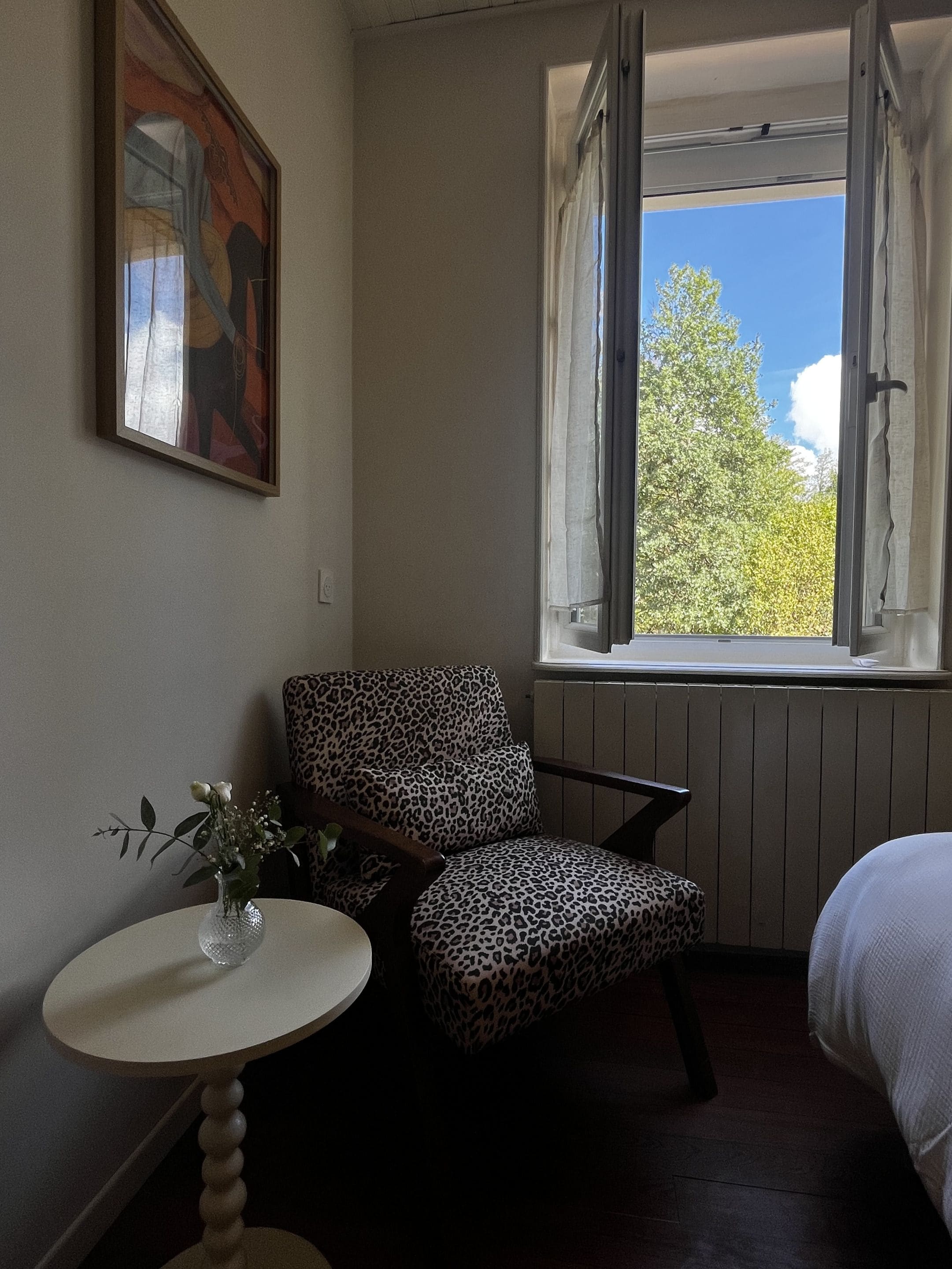 A room with a leopard-print chair, a small table with a vase, a painting on the wall, and a window with a view of trees.