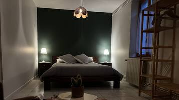 A bedroom with a bed, two bedside lamps, a hanging light fixture, a small potted plant, a patterned rug, and a wooden shelf unit.