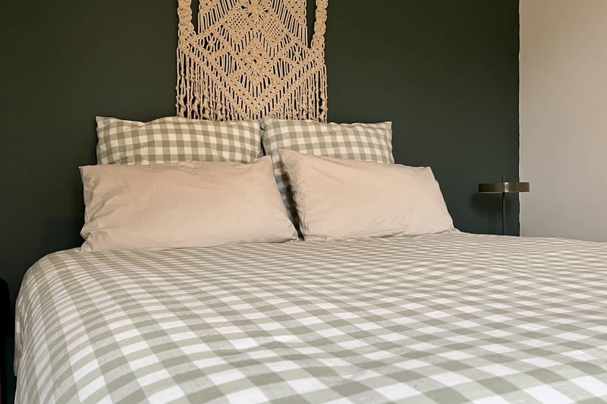 A bed with a checkered bedspread, two pillows, and a macramé wall hanging.