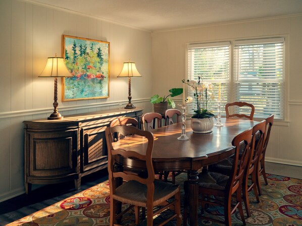 A dining room with a wooden table, six chairs, two lamps, a painting, a potted plant, and a window with blinds. - Masters Week Home Away from Home - 3BR Retreat Minutes from Augusta National (Augusta)