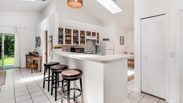 A modern kitchen with white cabinetry, a central island with stools, a dining area with a table and chairs, and a view of the outdoors through a glass door. - Stylish Retreat Near Beaches & Attractions - Private, Clean, Comfortable (Jacksonville)