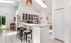 Une cuisine moderne avec des meubles blancs, un îlot central doté de tabourets, un coin repas avec une table et des chaises, et une vue sur l’extérieur grâce à une porte vitrée.