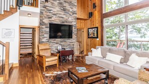 Amplia sala de estar con pared de piedra, escalera de madera, muebles de cuero y un ventanal con vista a un lago.