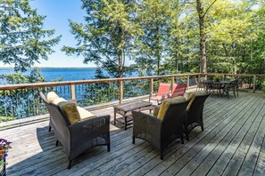 Une terrasse en bois, meublée de meubles en osier, qui offre une vue sur une étendue d’eau et est entourée d’arbres.