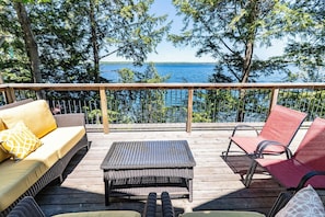 Une terrasse en bois avec un canapé, des chaises et une table, donnant sur une étendue d’eau et des arbres.