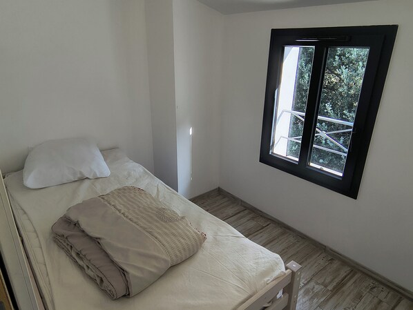 A single bed with pillows and a folded blanket, a window with a view of greenery, and a wooden floor. - New house on Oléron, 10 minutes from the beach (Le Grand-Village-Plage)
