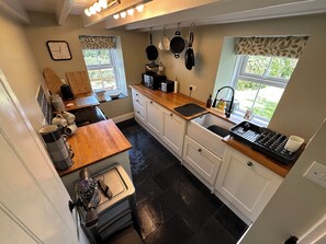 A compact kitchen with white cabinets, a wooden countertop, and a black tiled floor. There are two windows with leaf-patterned curtains, a sink with a black faucet, and a small window with a view of greenery outside. - Gorge View Welsh Slate Cottage (Cilgerran)
