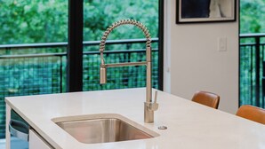 Cocina moderna con un lavamanos y grifo de acero inoxidable, encimera blanca y vistas a la vegetación a través de amplias ventanas.