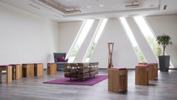 Ruime kamer met een hoge plafond, dakramen en een groot raam dat natuurlijk licht binnenlaat. Er staan houten banken en een unieke houten constructie in het midden.
