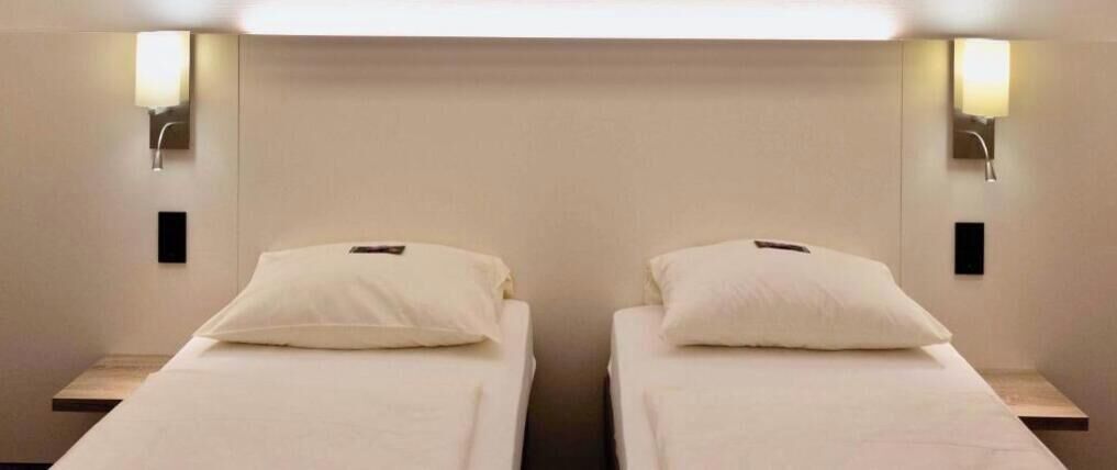 Two single beds with white linens and pillows, each with a bedside table and a wall-mounted lamp.