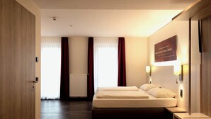 Een hotelkamer met een bed, een nachtkastje, een aan de muur bevestigde lamp en een raam met gordijnen.